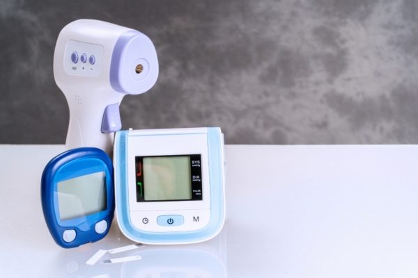 Sada domácich diagnostických prístrojov – bezkontaktný teplomer, glukomer s prúžkami a digitálny tlakomer na svetlom podklade.