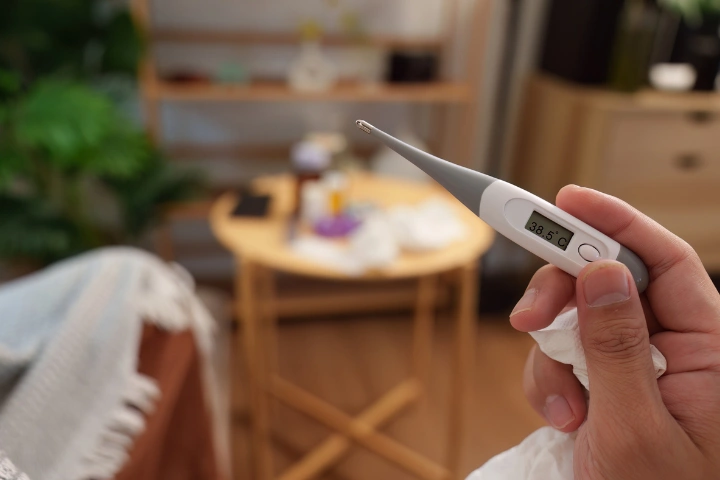 Ruka držiaca digitálny teplomer s nameranou hodnotou zvýšenej teploty v domácom prostredí.