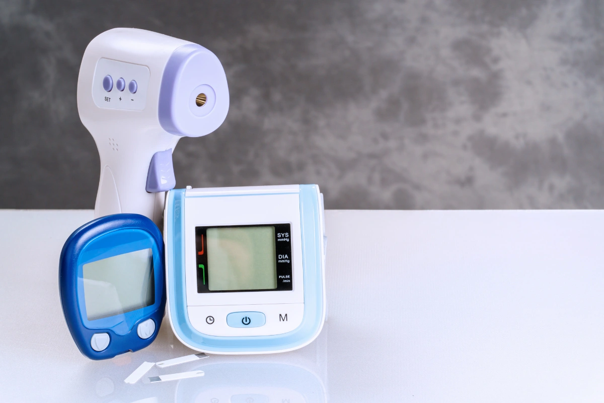Sada domácich diagnostických prístrojov – bezkontaktný teplomer, glukomer s prúžkami a digitálny tlakomer na svetlom podklade.