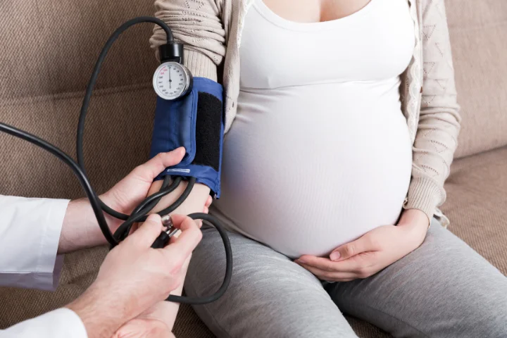 Těhotná žena sedí na gauči, zatímco jí zdravotnický pracovník měří krevní tlak.