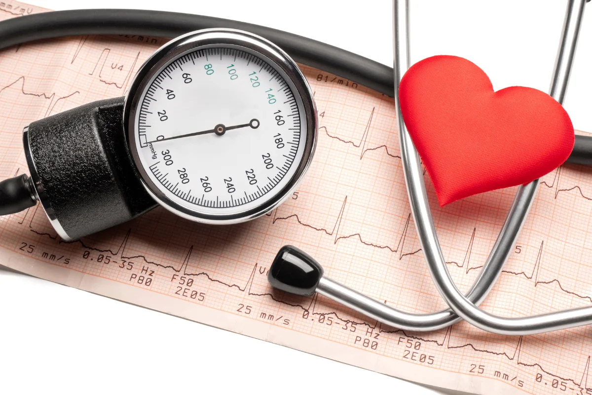 Mechanický tlakoměr na měření krevního tlaku položený na EKG záznamu vedle stetoskopu s červeným srdcem.