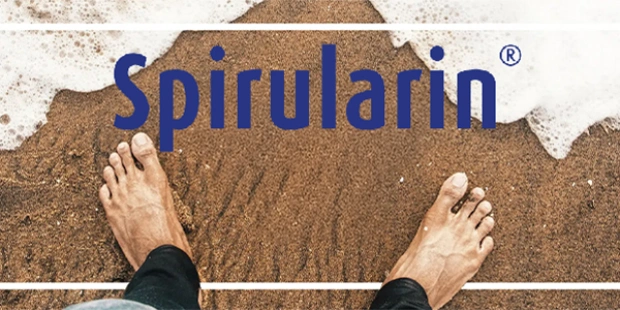 Modré logo Spirularin s pohľadom zhora na bosé nohy stojace na piesočnatej pláži.