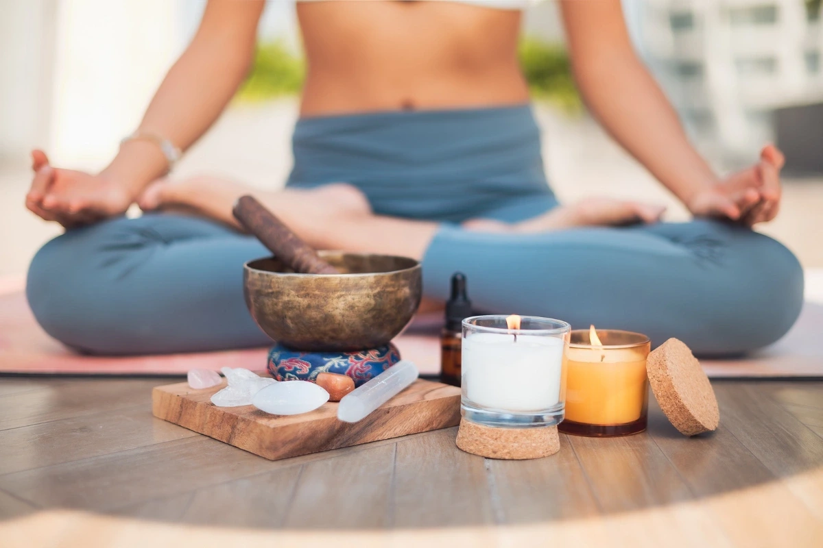 Žena meditujúca v pozícii lotosu s meditačnou miskou, krištáľmi a dvoma horiacimi sviečkami v popredí.