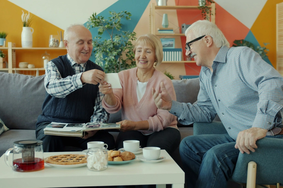 Skupina seniorov sa veselo rozpráva pri čaji a prezeraní fotografií, čo znázorňuje sociálnu inklúziu vďaka lepšiemu sluchu.